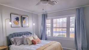 A bedroom with a bed, two framed artworks, a ceiling fan, and a window with curtains. - Hot Tub- Rose District-Walk to Shopping & Dining (Broken Arrow)