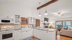 Een moderne keuken met witte kasten, een ingebouwde oven, magnetron en een groot eiland met een spoelbak. Er is een plafondventilator, hanglampen en uitzicht op de omgeving door een groot raam.