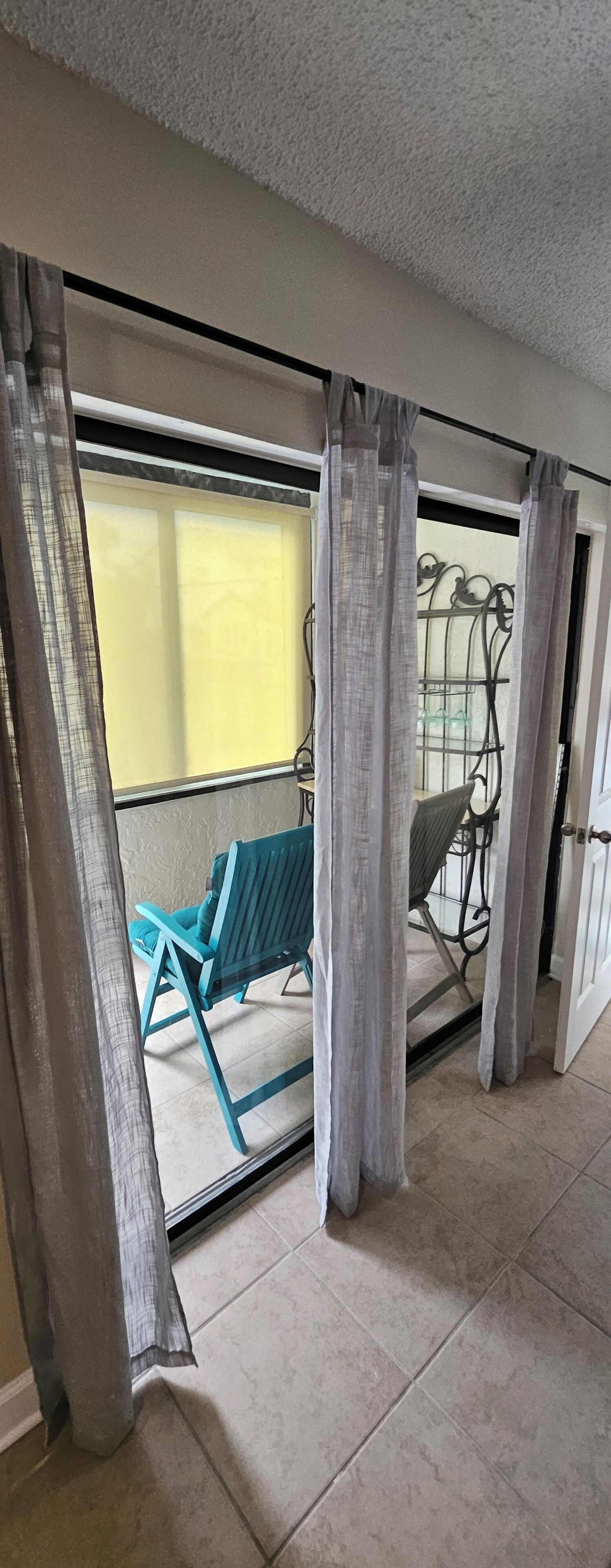 En balkon med en blå stol og en metalstol, gardiner trukket delvist til side, og udsigt til en gul mur og en dør.