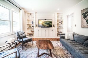 A modern living room with a flat-screen TV, wooden cabinets, and a cozy seating area. - Woodstock Family Home w/ Tree House + Heated Pool (Woodstock)