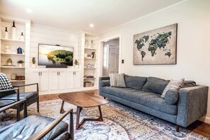 A living room with a grey sofa, a coffee table, and a TV on a white cabinet. There is a world map on the wall and shelves with various items. - Woodstock Family Home w/ Tree House + Heated Pool (Woodstock)