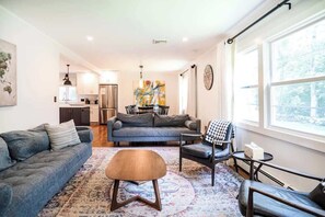Un salon moderne avec un canapé, un fauteuil et une table basse, une cuisine en arrière-plan et une grande fenêtre donnant sur un beau paysage.