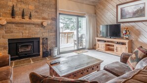 A cozy living room with a stone fireplace, wooden furniture, a television, and a scenic view through the glass door. - Snowcreek #309  2 Plus Loft, 2 Bathroom Condo Sleeps 8 People with Amazing Mountain Views (Mammoth Lakes)