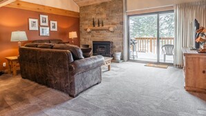 A cozy living room with a fireplace, a leather sofa, and a large window overlooking a deck. - Snowcreek #309  2 Plus Loft, 2 Bathroom Condo Sleeps 8 People with Amazing Mountain Views (Mammoth Lakes)