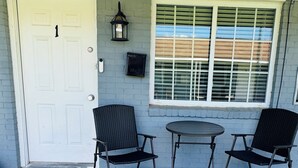 A front porch with a white door, two wicker chairs, a small round table, and a window with blinds.