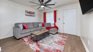A living room with a grey sectional sofa, a wooden coffee table, a red and white rug, a ceiling fan, and a white door.