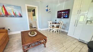 Een woonkamer met een houten salontafel, een aan de muur bevestigde televisie en een eethoek met een witte tafel en stoelen.