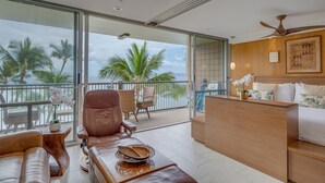 A modern hotel room with a balcony, leather seating, a wooden desk, and a bed with tropical-themed bedding. - Stunning retreat with oceanfront views (Kihei)