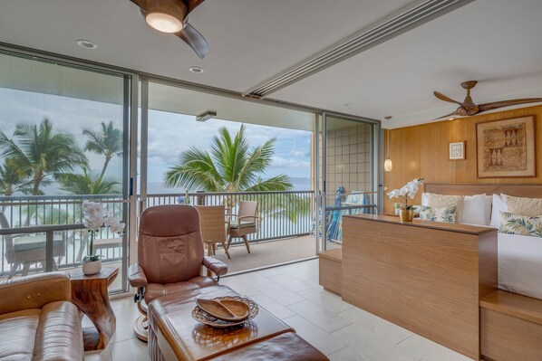 A modern hotel room with a balcony, leather seating, a wooden desk, and a bed with tropical-themed bedding.