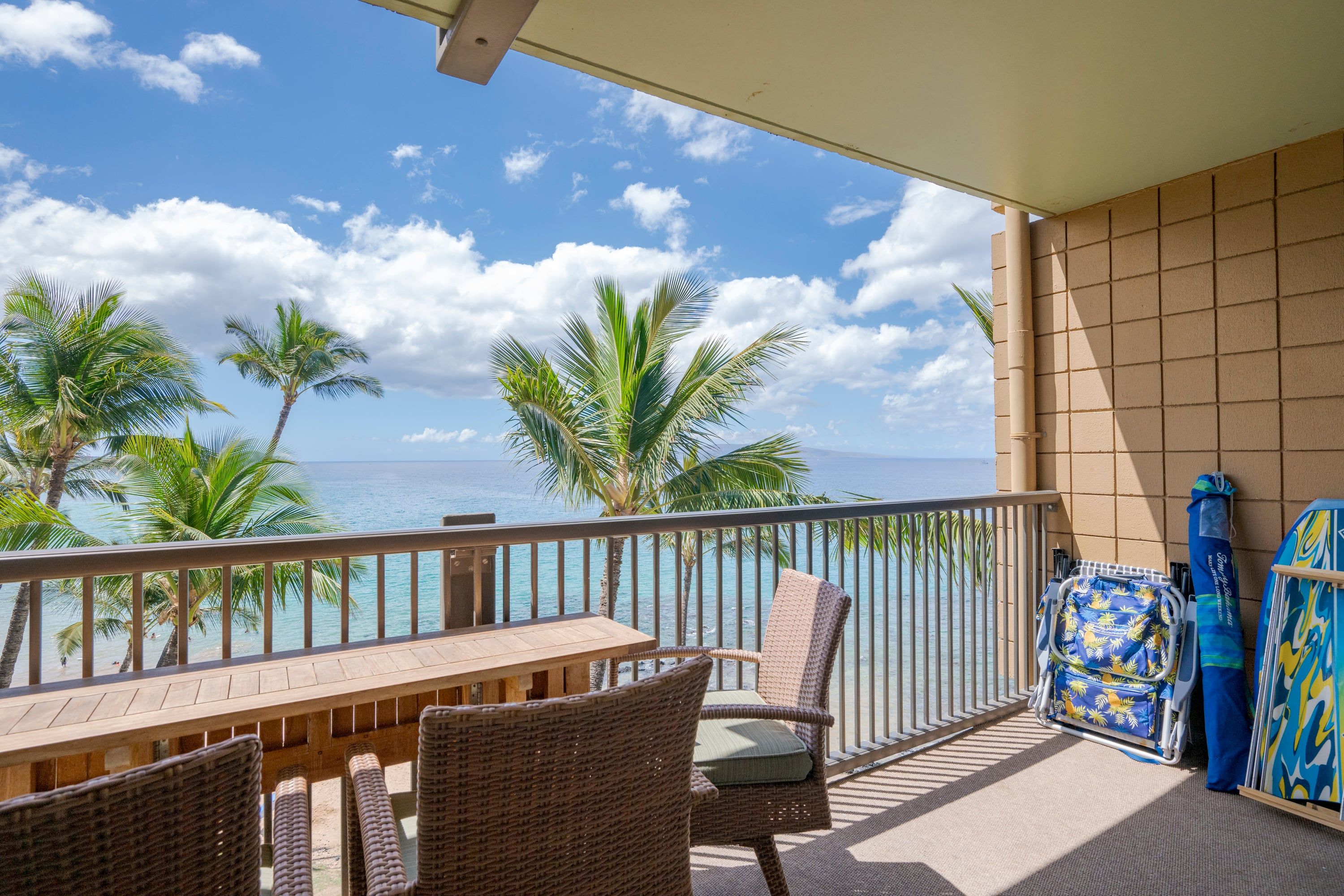 A balcony with a wooden table and wicker chairs, overlooking a beach with palm trees and the ocean.