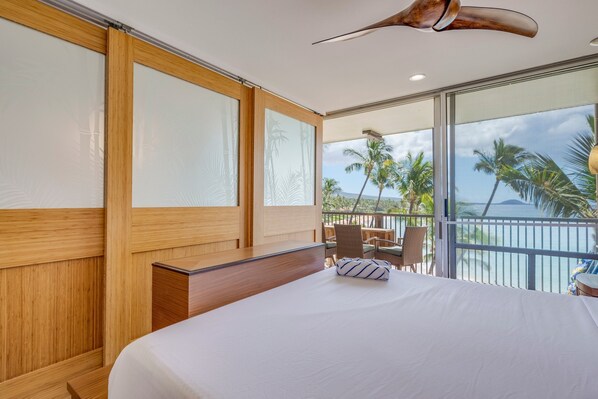 A hotel room with a large bed, wooden paneling, a desk, and a balcony with a view of palm trees and the ocean.