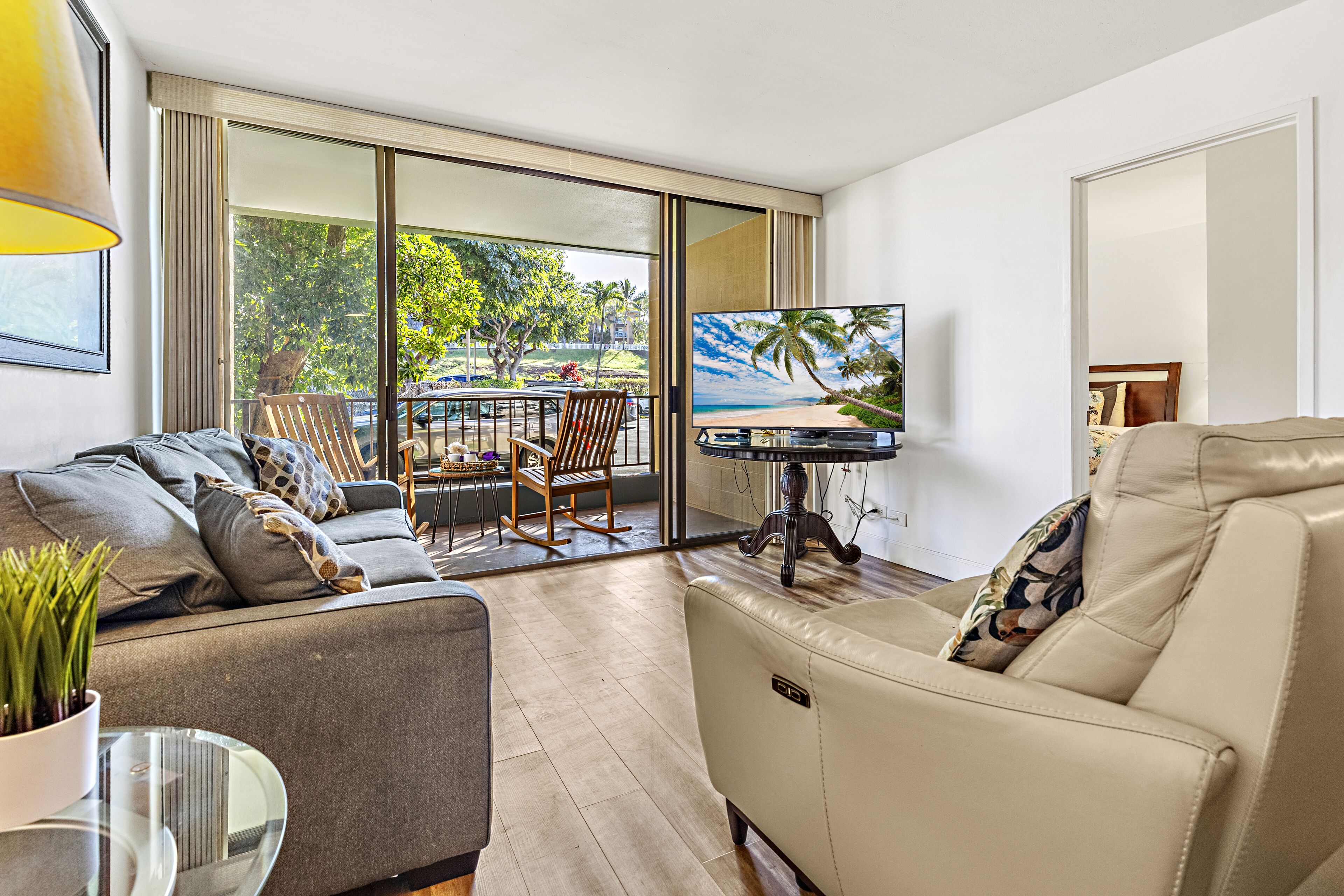 A modern living room with a sofa, a chair, a TV, and a balcony with a view of trees and a beach.