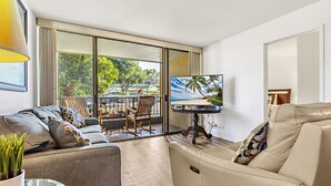 Una sala de estar moderna con un sofá, una silla, un televisor y un balcón con vistas a árboles y una playa.