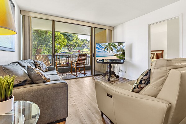 A modern living room with a sofa, a chair, a TV, and a balcony with a view of trees and a beach.