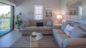 A living room with a sofa, a coffee table, and a lamp. There are two windows with blinds, a plant, and a wall-mounted mirror. - Ocean front condo,  walk to quaint Vero Beach restaurants and shops.  (Vero Beach)