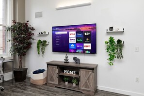 Una sala de estar con un televisor montado que muestra servicios de transmisión, un mueble bajo de madera con un letrero que dice 'HOME' y plantas en macetas.