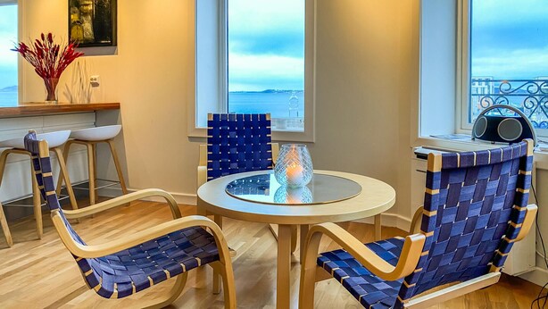 Ein modernes Zimmer mit einem runden Tisch, zwei Stühlen, einer Vase mit roten Zweigen und Blick auf das Meer durch große Fenster.