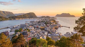 Eine Küstenlandschaft mit einer Mischung aus moderner und traditioneller Architektur, einem Yachthafen mit Booten und einem Bergpanorama im Hintergrund.