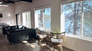 Ett modernt vardagsrum med en svart soffa, två fåtöljer i rotting och stora fönster som ger utsikt över träd och en sjö eller ett annat vattendrag.