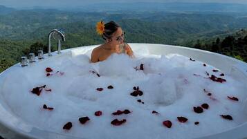 A person in a hot tub filled with water and rose petals, with a scenic view of a mountainous landscape in the background.