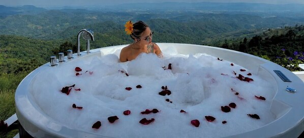 A person in a hot tub filled with water and rose petals, with a scenic view of a mountainous landscape in the background.