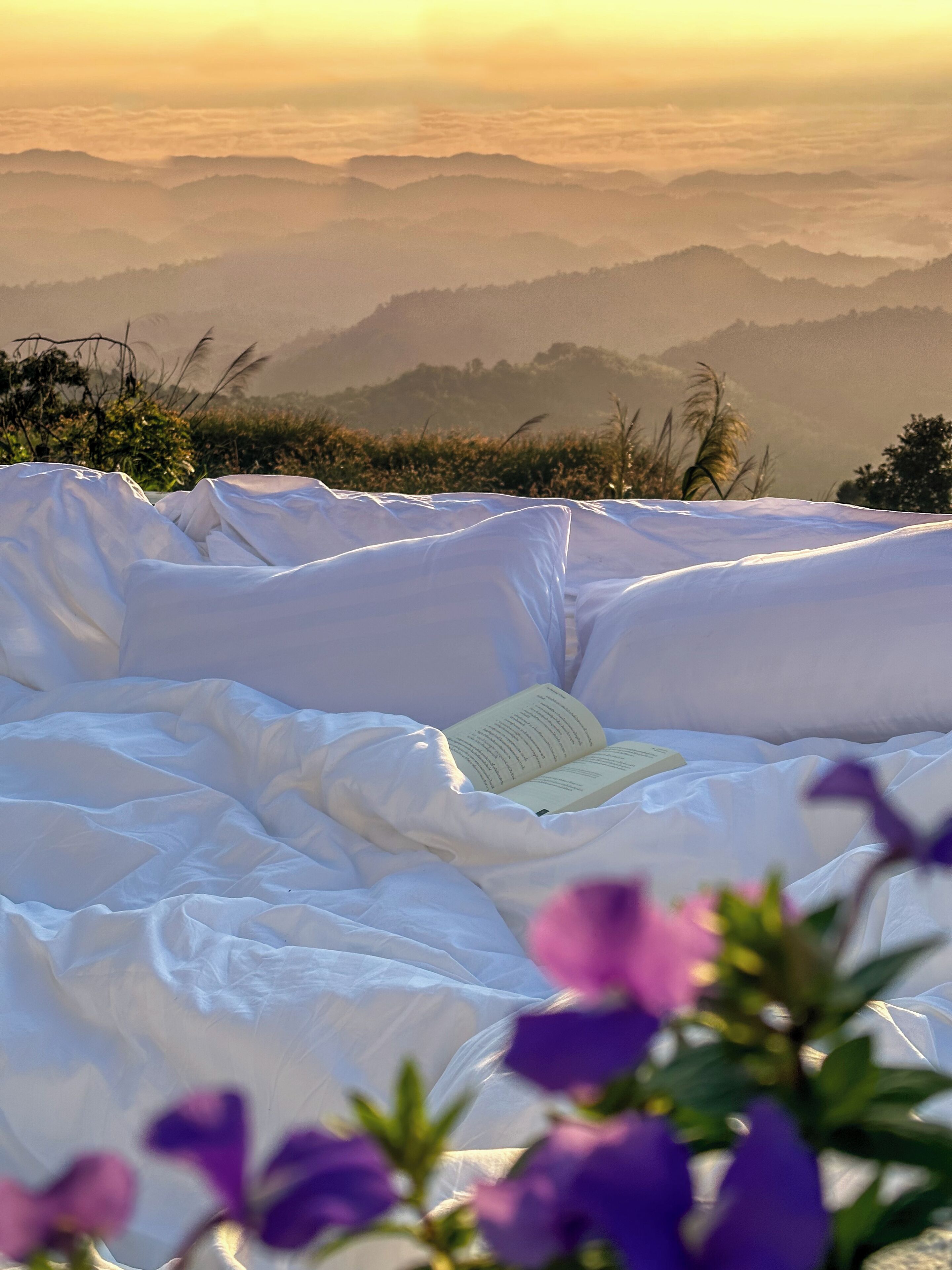 A bed with a book on it, surrounded by white bedding, with a view of distant mountains and some purple flowers in the foreground.
