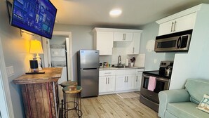 Una cocina compacta con armarios blancos, un refrigerador de acero inoxidable, un microondas y una estufa. Hay una barra con taburetes y un televisor montado en la pared.
