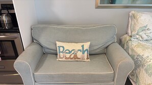 Un sofá gris claro con un cojín que tiene la palabra 'Beach' y una pequeña ilustración de una planta.