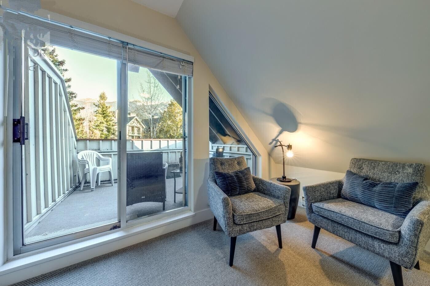 A cozy room with a large sliding glass door leading to an outdoor patio, two armchairs, a small side table with a lamp, and a sloped ceiling.