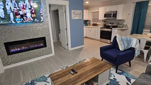 A modern living room with a flat-screen TV, a ceiling fan, a fireplace, and a kitchen in the background.