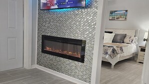 A modern living room with a flat-screen TV mounted above a fireplace, a bed with a floral bedspread, and a patterned rug.
