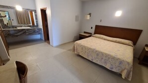 Un dormitorio con cama, mesita de noche, tocador, espejo y baño con lavabo y espejo.