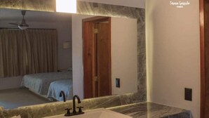 Un baño con lavamanos de mármol, ventilador de techo y espejo que refleja un dormitorio con cama y puerta.