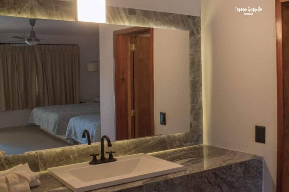 A bathroom with a marble sink, a ceiling fan, and a mirror reflecting a bedroom with a bed and a door.