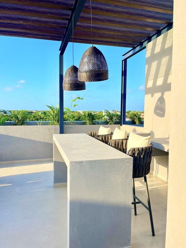 An outdoor seating area with a concrete table and chairs, two woven pendant lights, and a view of greenery and buildings in the distance.