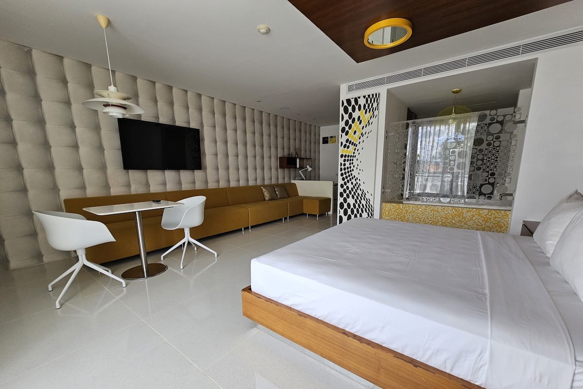 A modern bedroom with a large bed, a sofa, a dining table with chairs, and a TV mounted on the wall.