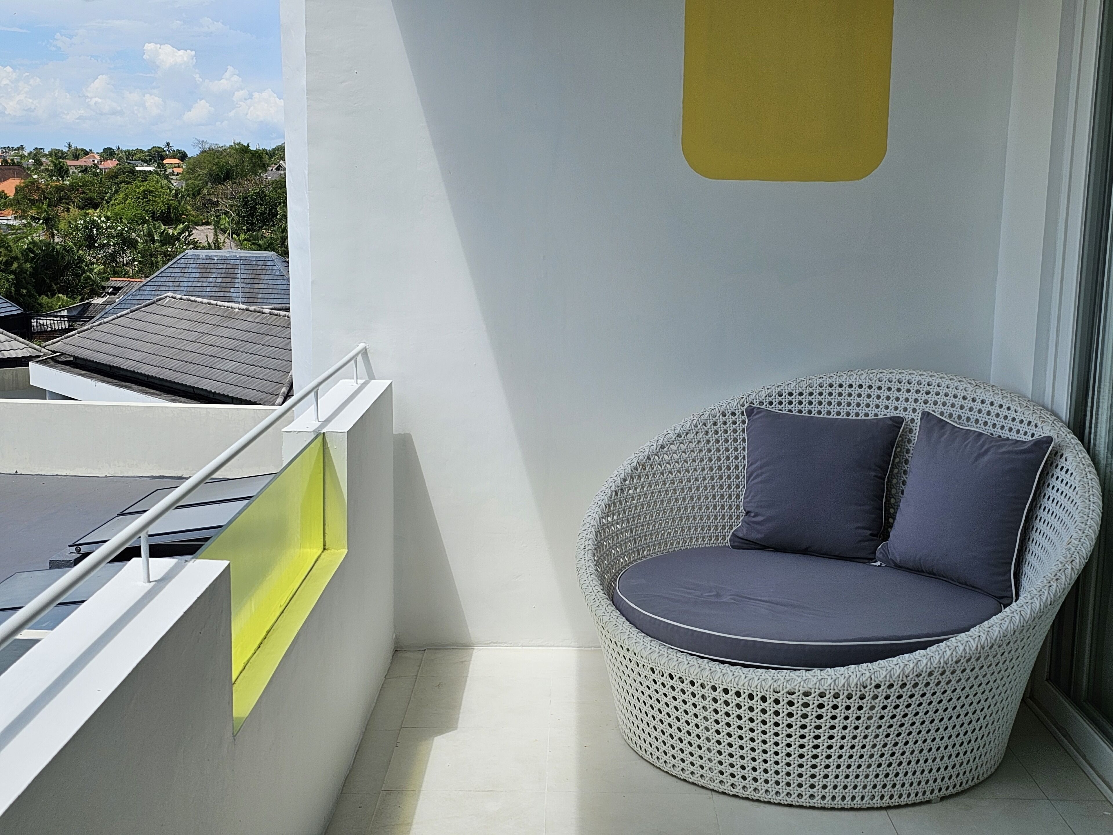 A balcony with a white wicker chair and cushions, a yellow window, and a view of houses in the background.