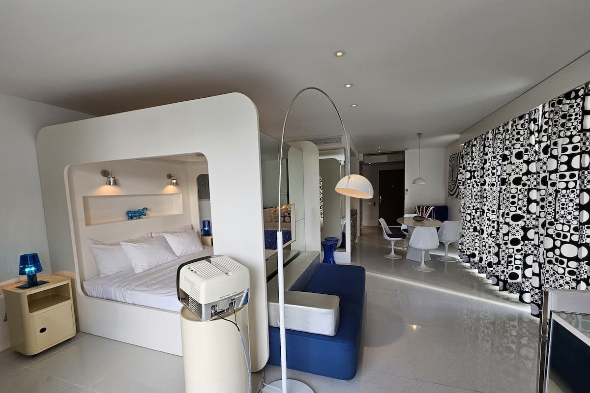 A modern hotel room with a bed, bedside table, lamp, and a large mirror. There is a dining area with a table and chairs, and a patterned wall.