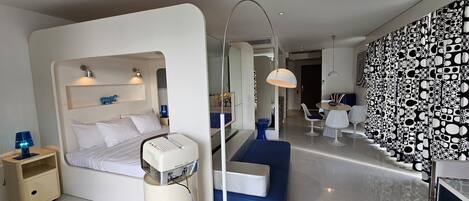 Une chambre d’hôtel moderne avec un lit, une table de chevet, une lampe et un grand miroir. On y trouve également un coin repas avec une table et des chaises, ainsi qu’un mur décoré de motifs.