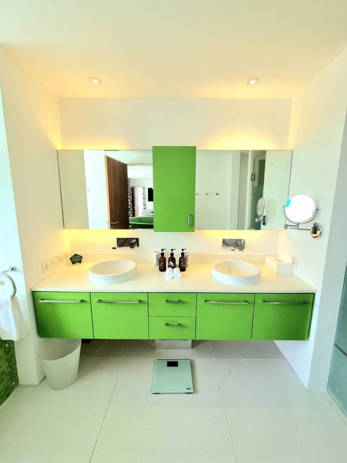 Une salle de bain moderne avec deux lavabos, des meubles verts, une balance et un grand miroir.