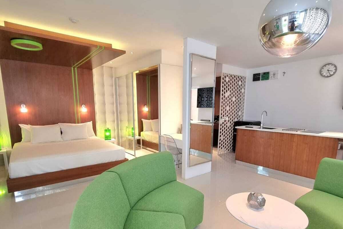 A modern hotel room with a curved green sofa, a small round table, a bed with white linens, a wooden headboard, and a kitchen area with a sink and countertop.