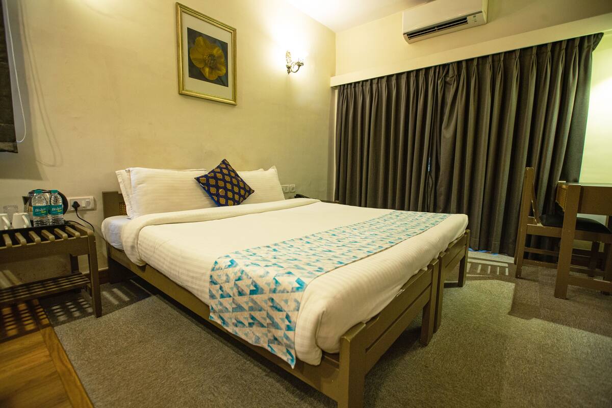 A hotel room with a bed, a bedside table with a lamp, a chair, and a window with curtains.