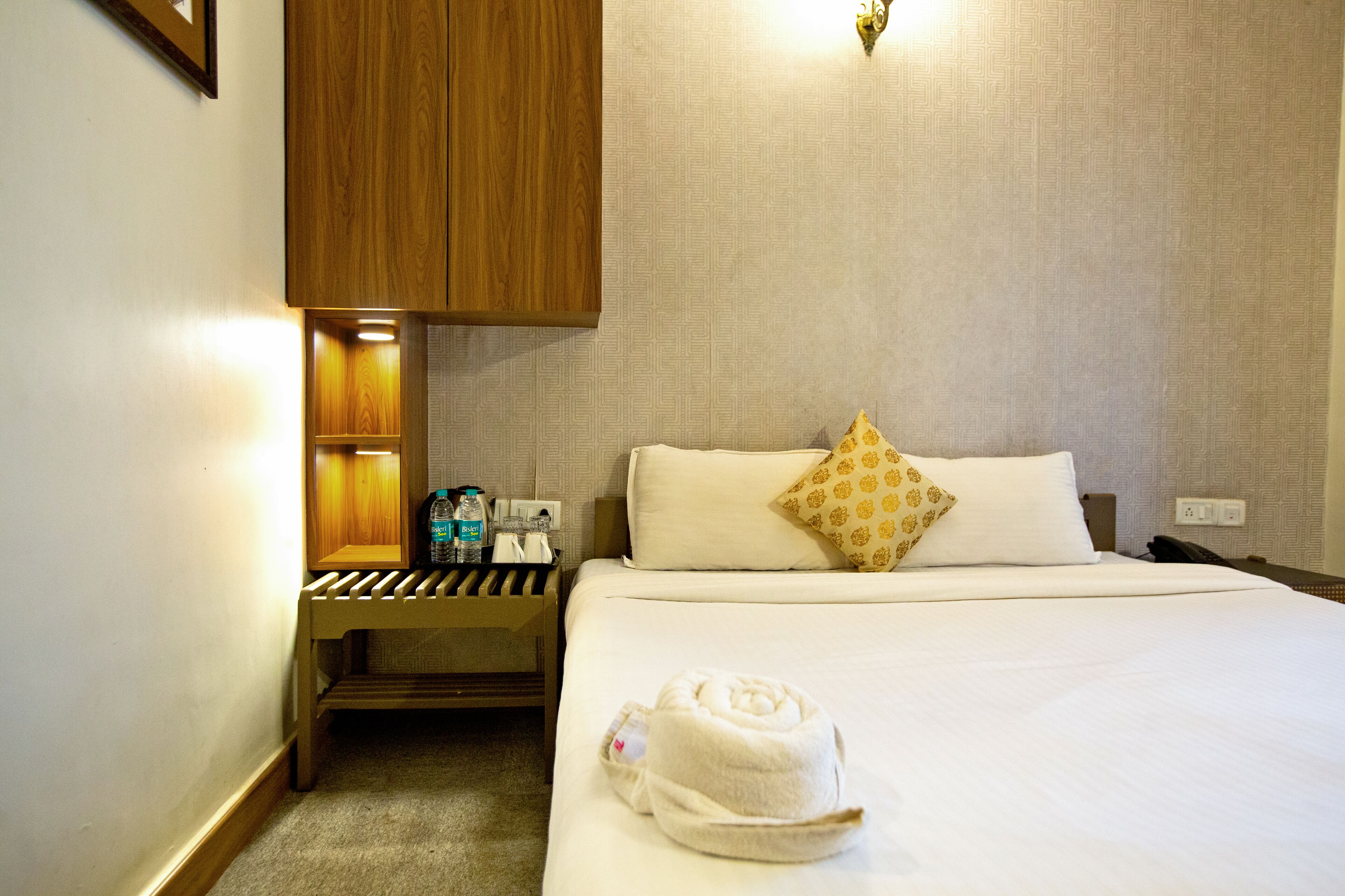 A hotel room with a bed, a nightstand, a wooden cabinet, and a wall-mounted light fixture.