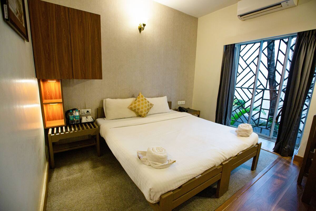 A hotel room with a bed, bedside table, and a window with a view of greenery.