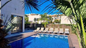 Una piscina con sillones de descanso, rodeada de palmeras y edificios modernos.