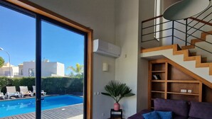 Una sala moderna con un amplio ventanal corredizo que da a una piscina, un sofá morado, una mesita auxiliar con una planta y una escalera de madera.