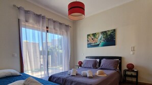 Habitación de hotel con dos camas, un ventanal con cortinas traslúcidas, un cuadro de una escena tropical y una lámpara colgante roja.