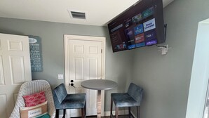 Una habitación con una puerta blanca, un televisor montado mostrando varios canales, dos sillas azules, una mesita redonda y una silla de mimbre con un cojín que dice 'playa'.