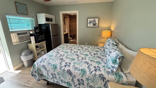 Un dormitorio con cama, ventilador de techo, lámpara, microondas, refrigerador, ventana con persianas y puerta que da a un baño.