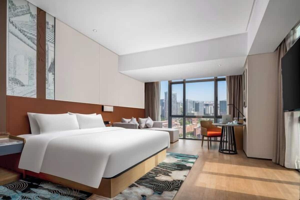 A modern hotel room with a large bed, a desk, a chair, a view of the city, and a large window.